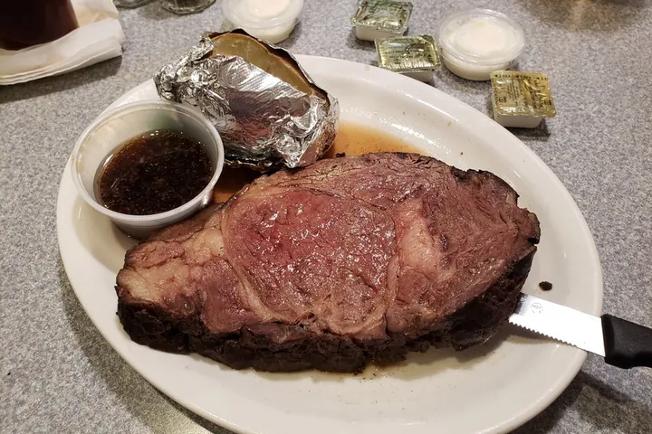 A cut of prime rib with a baked potato and a cup of au jus